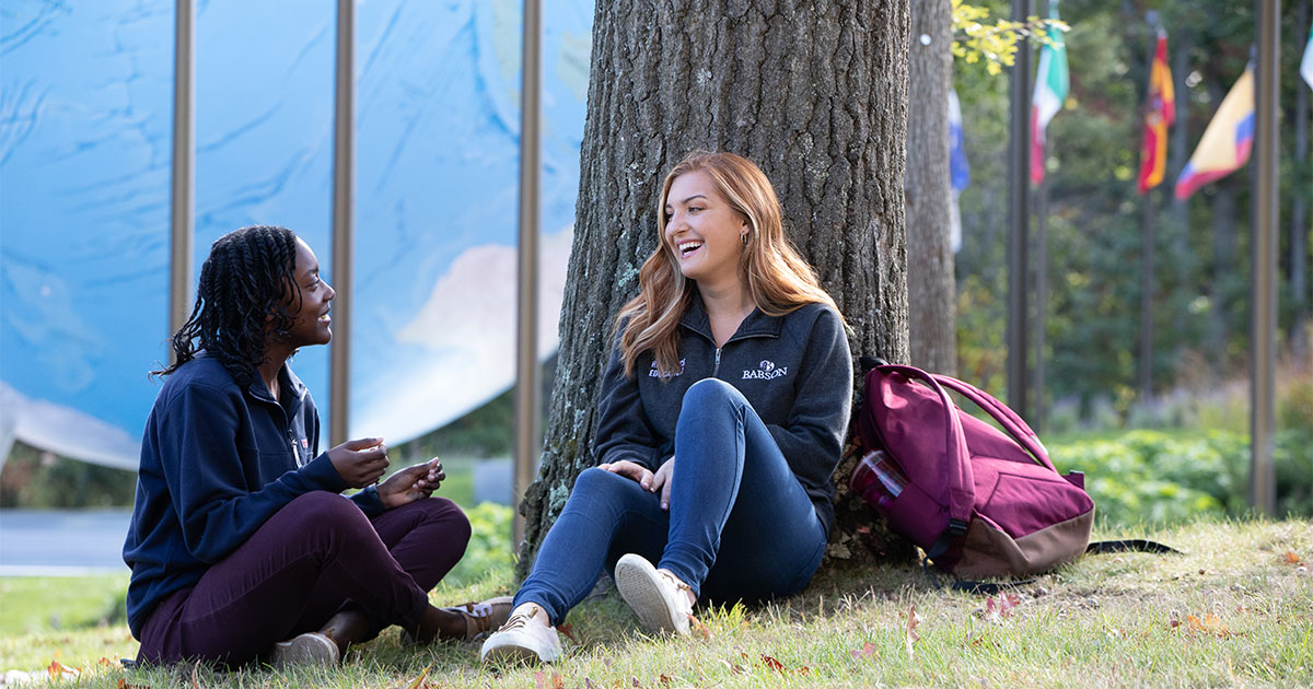 Two students sit under a tree near the Babson Globe