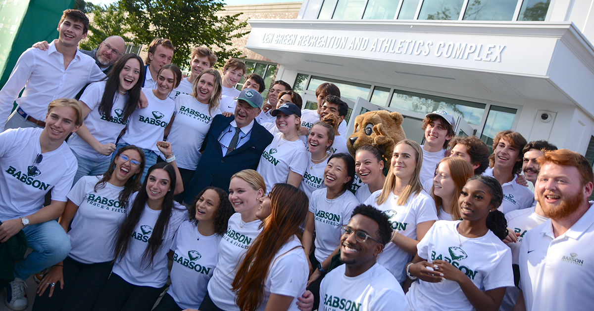 A group of student-athletes in white T-shirts pose for a photo with Len Green in front of the building named for him