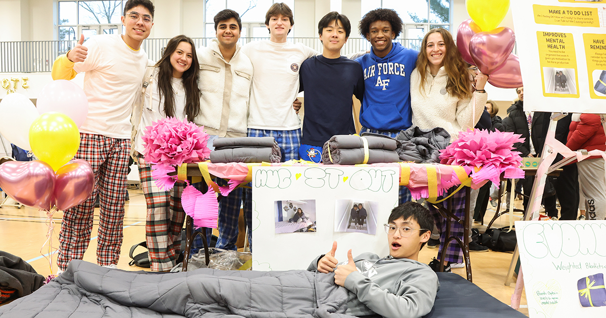 One student lies under a weighted blanket in front of a group of students posing for a photo