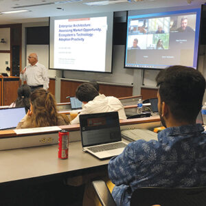 Students on laptops watch a professor inn front a large screen of online students