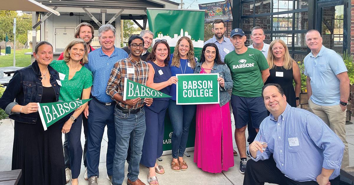 A group of alumni pose for a photo holding Babson banners and pennants
