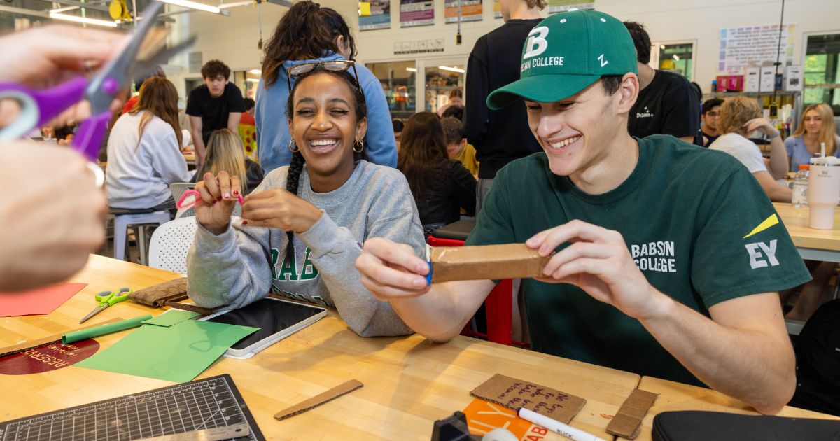 Two students work on projects involving paper and cardboard in a boisterous classroom