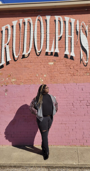 Sevyn Williams poses for a photo while leaning against a wall that reads Rudolph's