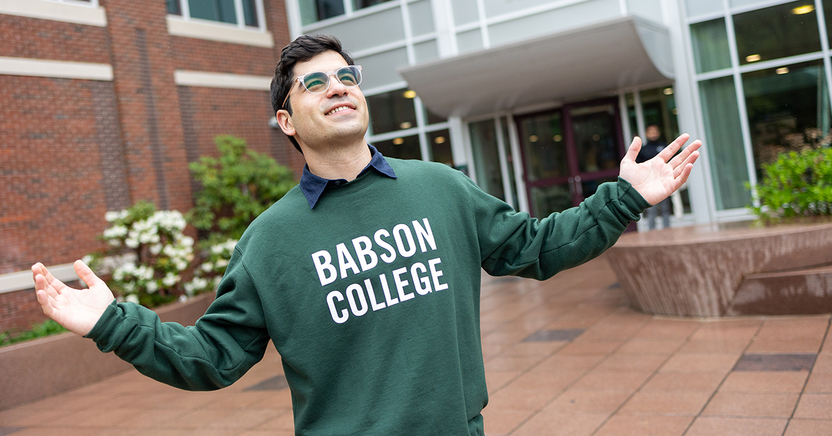 In a Babson sweatshirt and with his arms up, Robert Pulles poses for a photo outside Olin Hall