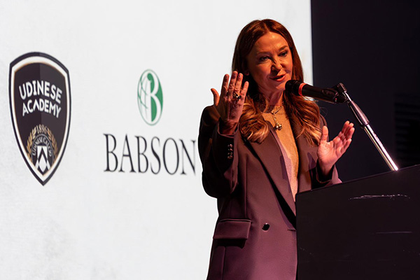 A woman speaks at a podium in front of a backgroup with the logos of Babson and Udinese Calcio