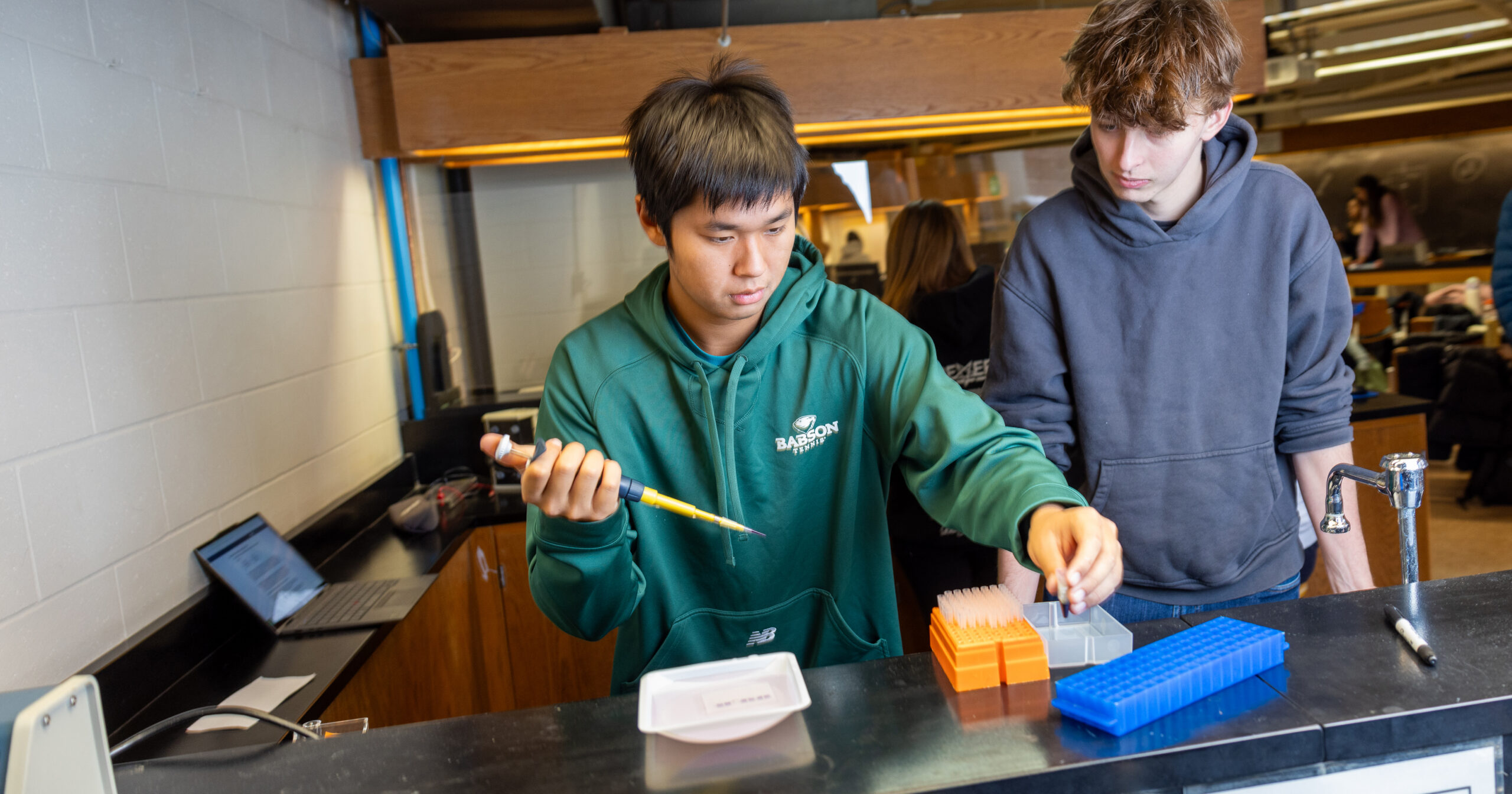 Two students work together at a laboratory bench. One student uses a pipette to transfer liquid into a small container, while the other stands beside them observing. The workspace includes trays of plastic test tubes, a laptop, and other lab equipment. The setting appears to be a science classroom or lab with additional students visible in the background.