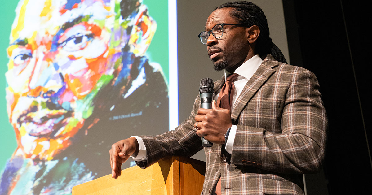 Standing on a stage, Derron Wallace speaks on a microphone near an image of Martin Luther King Jr.