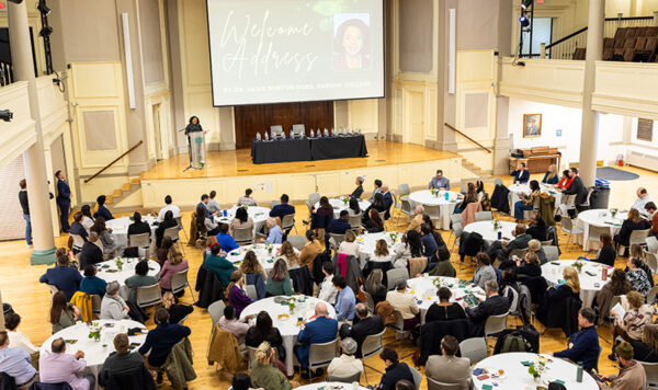 An overview of the room packed with tables while Sadie Burton-Goss speaks at a podium on a stage