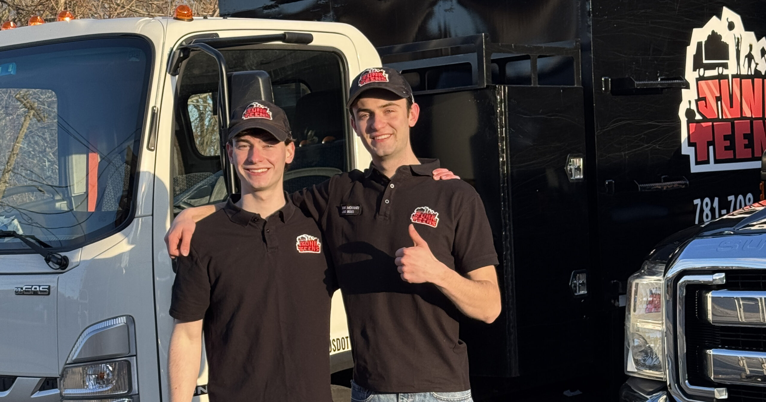 Two people wearing matching black polos and caps with a red-and-white logo stand side by side in daylight in front of a white cab truck and a black box truck; one person has an arm around the other’s shoulder, and the black truck shows the logo and a partially visible phone number starting with “781.”