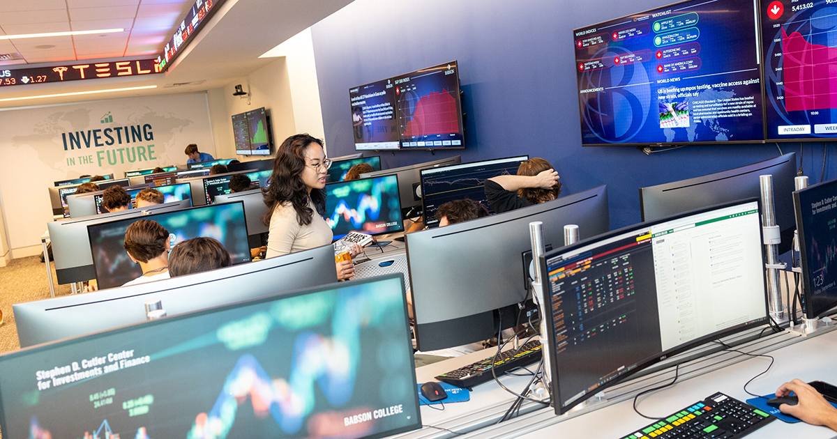 A female student looks at a computer screen in a finance lab filled with computers and a scrolling stock ticker.