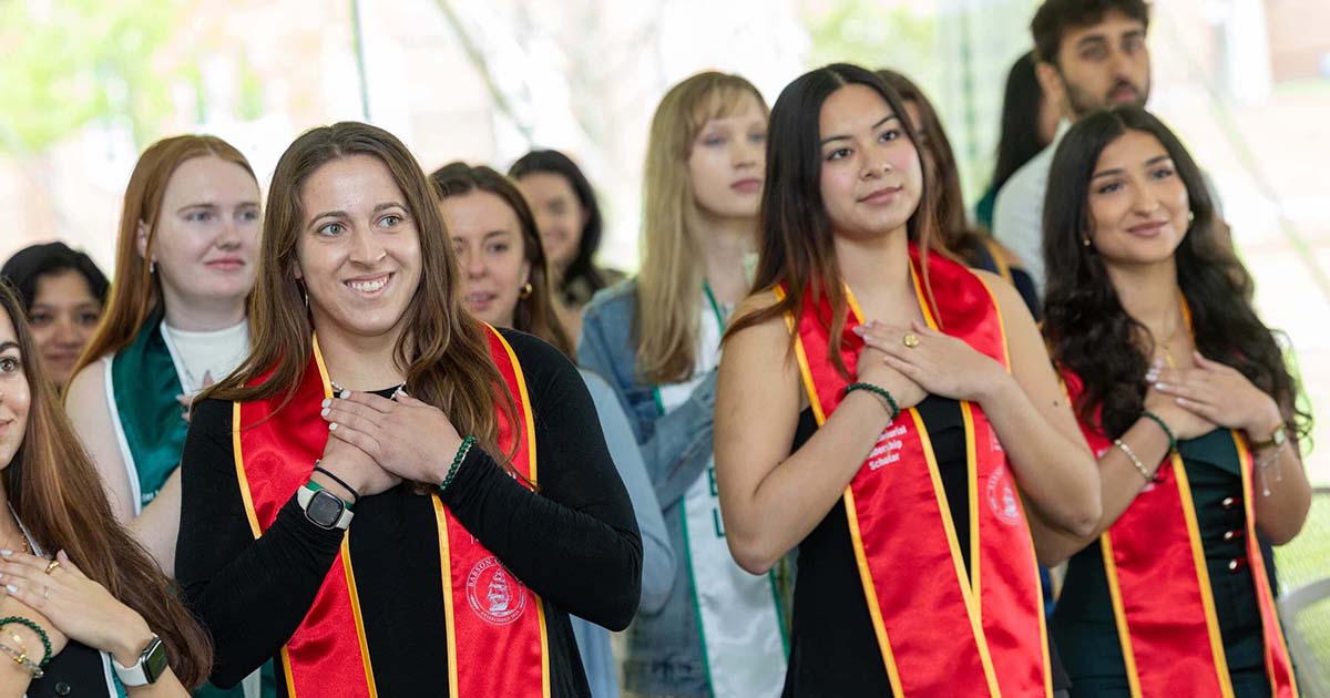 Graduates wearing their stoles stand while placing their hands over their hearts