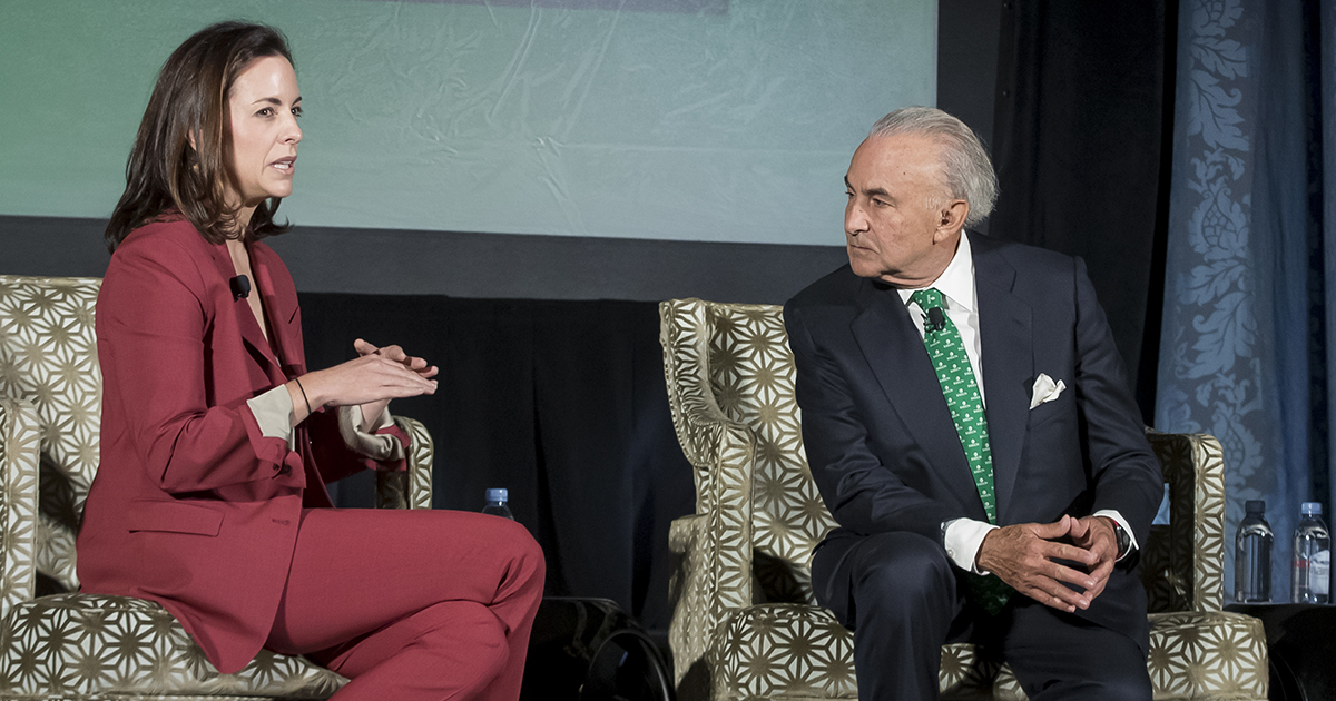 Adriana Cisneros speaks while sitting on a stage next to Gustavo Cisneros