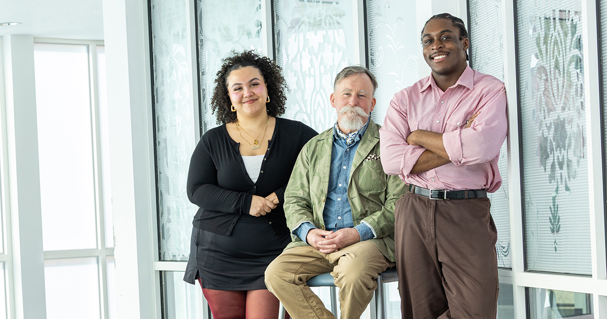 Jolie Wyatt, Rick Blackshaw, and Kanayoliseh Onwudiegwu pose for a portrait with Rick sitting between the two students, who are standing