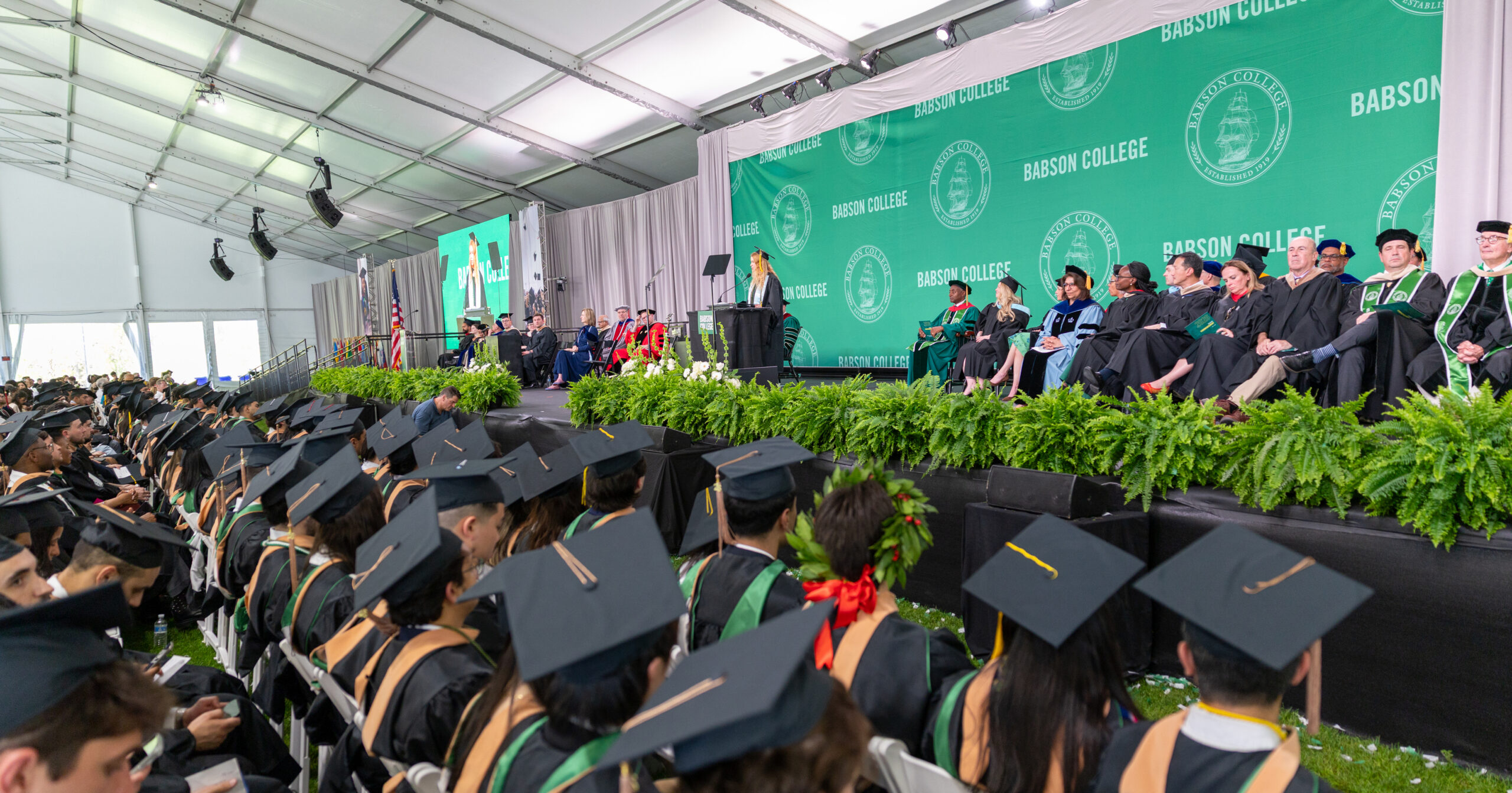 Wide-angle photo of a graduation ceremony inside a large white event tent at Babson College. In the foreground, rows of graduates wearing black caps and gowns with green accents sit facing a raised stage. Some graduates have decorative caps or accessories. The stage runs across the right side of the image and is lined with green ferns and floral arrangements. On the stage, a group of officials in academic regalia sit in a row, while one speaker stands at a podium delivering remarks. Behind the stage is a large green backdrop repeatedly printed with “BABSON COLLEGE” and the college seal, along with a large screen displaying the speaker. An American flag stands near the stage, and lighting and sound equipment hang from the tent ceiling above.