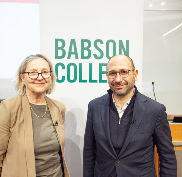 Smiling woman and man stand in room with Babson College sign.
