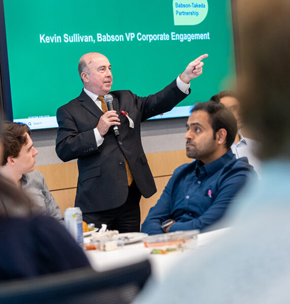 Man in suit gestures while speaking to a classroom of people.
