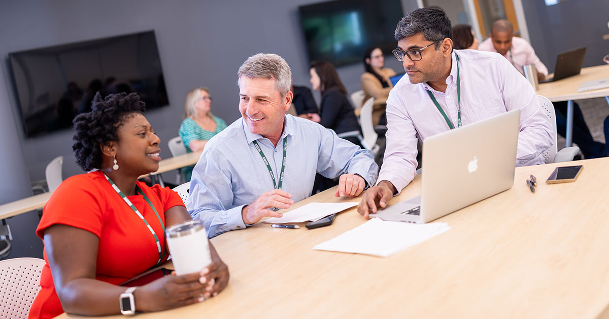 Three people sit around a table, looking at a laptop