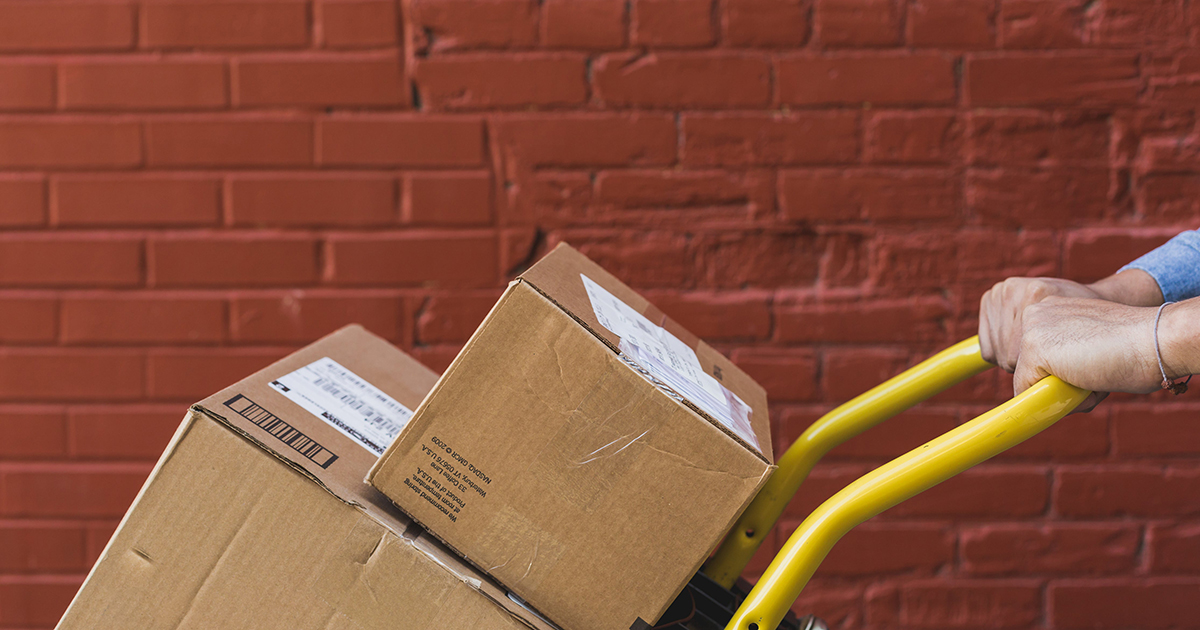 Arms pushing yellow trolley packed with delivery boxes.