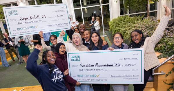 A group of students celebrate while holding up two giant checks