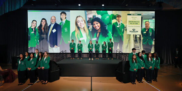 Students in green jackets stand on stage in front large images of students