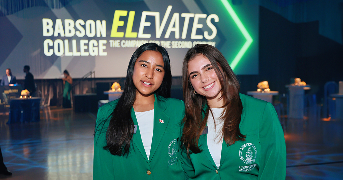 Two students in green jackets pose for a photo in front of the Babson Elevates logo