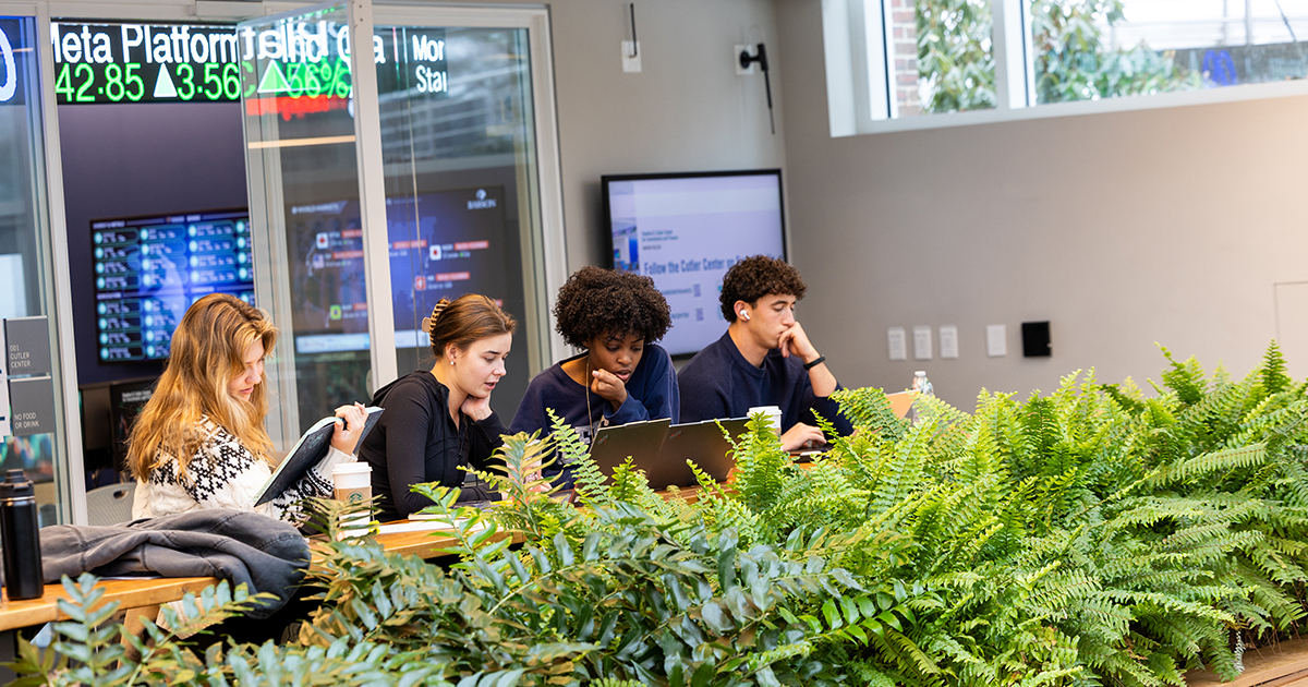 A line of student sit at a long table while working on their laptops
