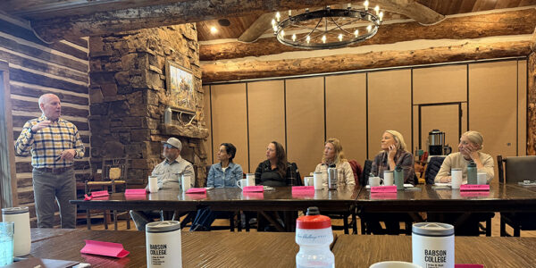 A professor stands at the left of a long table, where participants sit and listen
