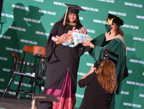 A student, holding her child, poses for a photo while crossing the commencement stage.